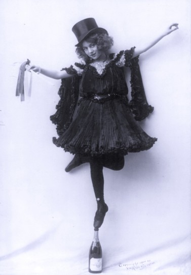Model posed in ornate costumes: in black pressed pleats, with top hat; standing tip-toe on champagne bottle